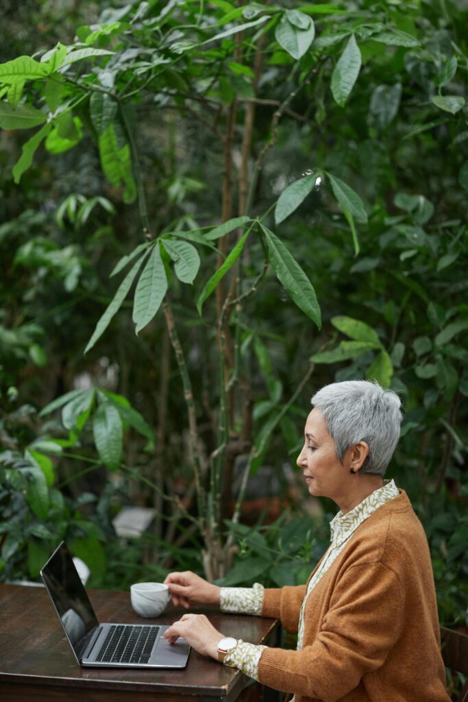 pexels-photo-6787887-6787887 Elderly woman using a laptop in a lush garden, holding a coffee cup.